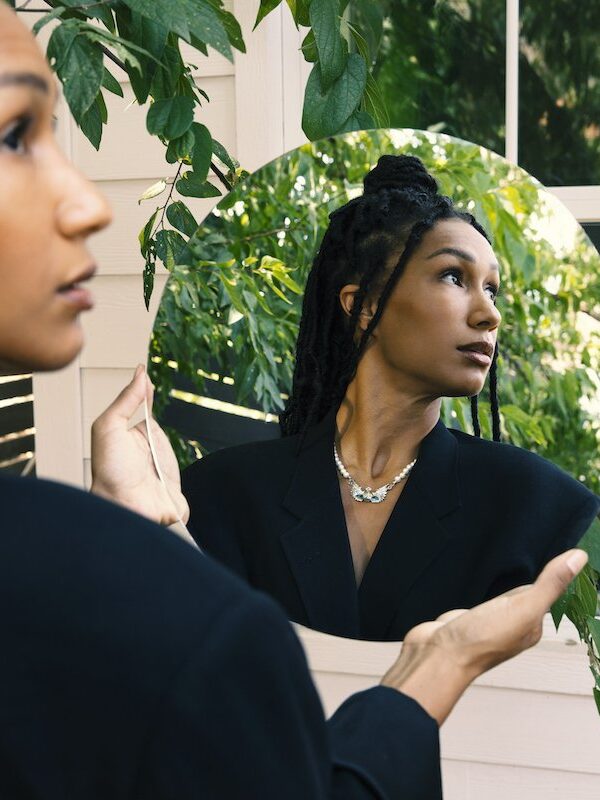 A person holding a circular mirror outdoors, reflecting their face and upper body with greenery and a window in the background.