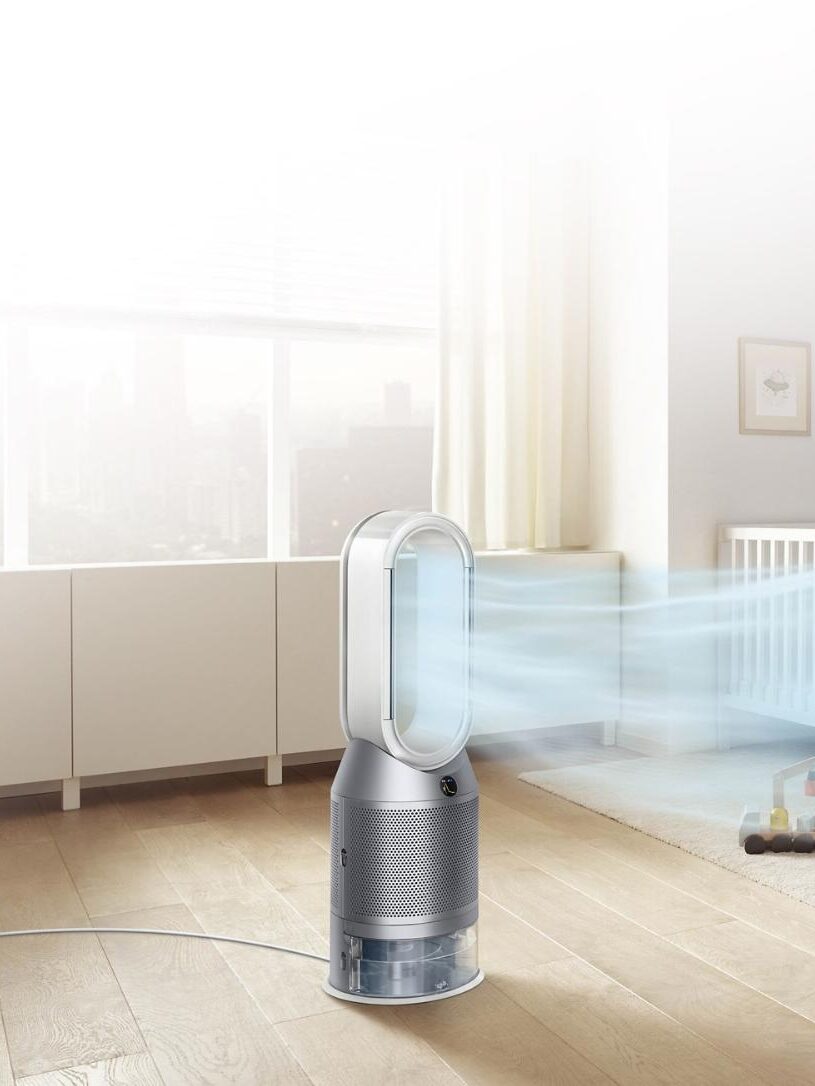 A woman sits on a chair, reading to a child in a bright room with a crib and toys. A bladeless fan is running, emitting a visible airflow.