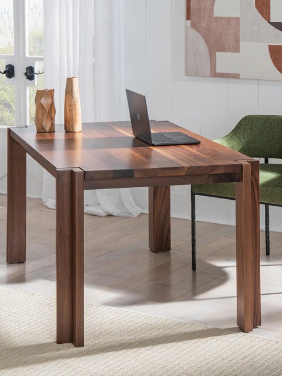 A wooden desk with a laptop and wooden vase decor stands in a bright room. A green upholstered chair is beside the desk, and an abstract art piece hangs on the wall.