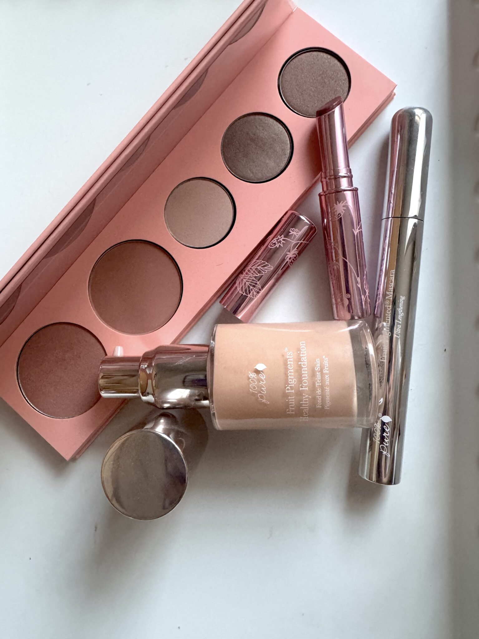 A collection of makeup items including a pink eyeshadow palette, foundation, two lip glosses, and a tube of mascara arranged on a white surface.