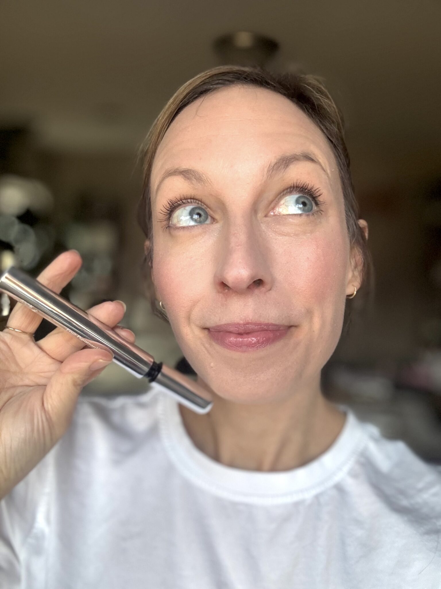 A woman in a white shirt holds a silver makeup product near her face and looks upward with a thoughtful expression.