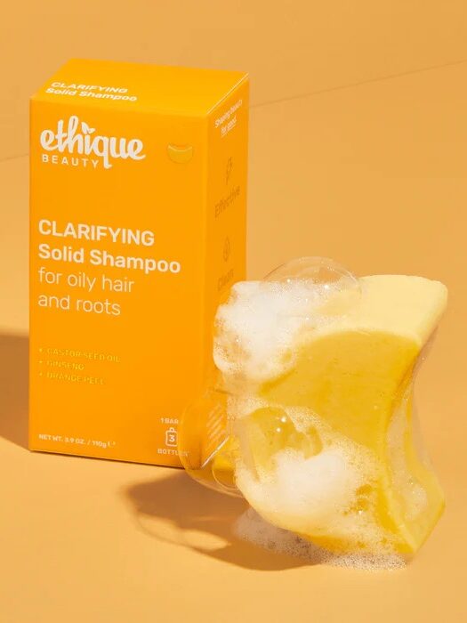 A yellow box labeled "Ethique Beauty Clarifying Solid Shampoo" is next to a foamy yellow shampoo bar on an orange background.