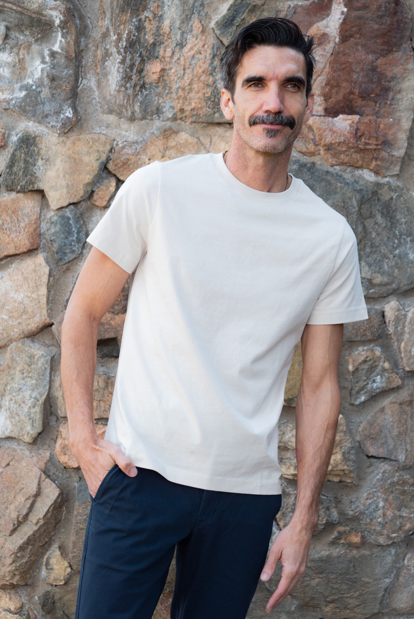 A man with short dark hair and a mustache stands in front of a stone wall, wearing a plain white t-shirt and dark pants.