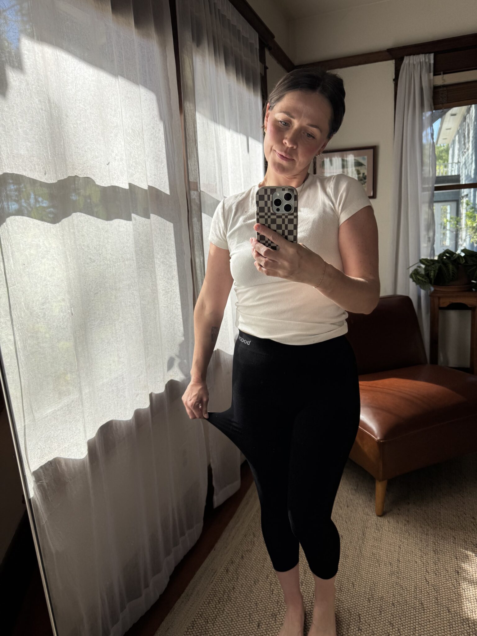 Woman stands indoors in natural light, wearing a white shirt and black leggings, holding out waistband and taking a mirror selfie with a phone.