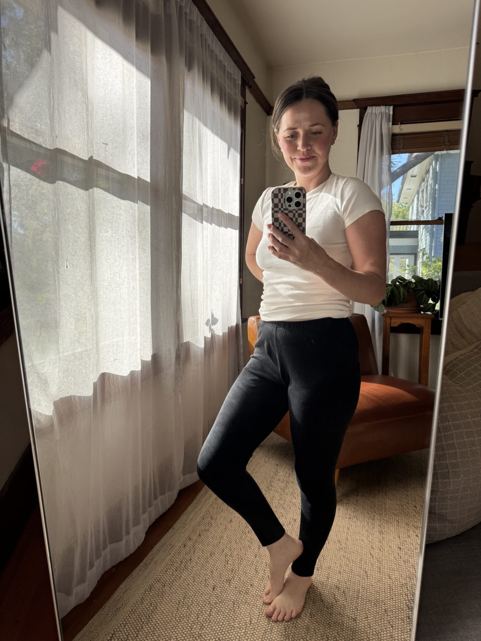 A person stands barefoot indoors, wearing a white t-shirt and black leggings, taking a mirror selfie in a sunlit room with sheer curtains and a brown chair.