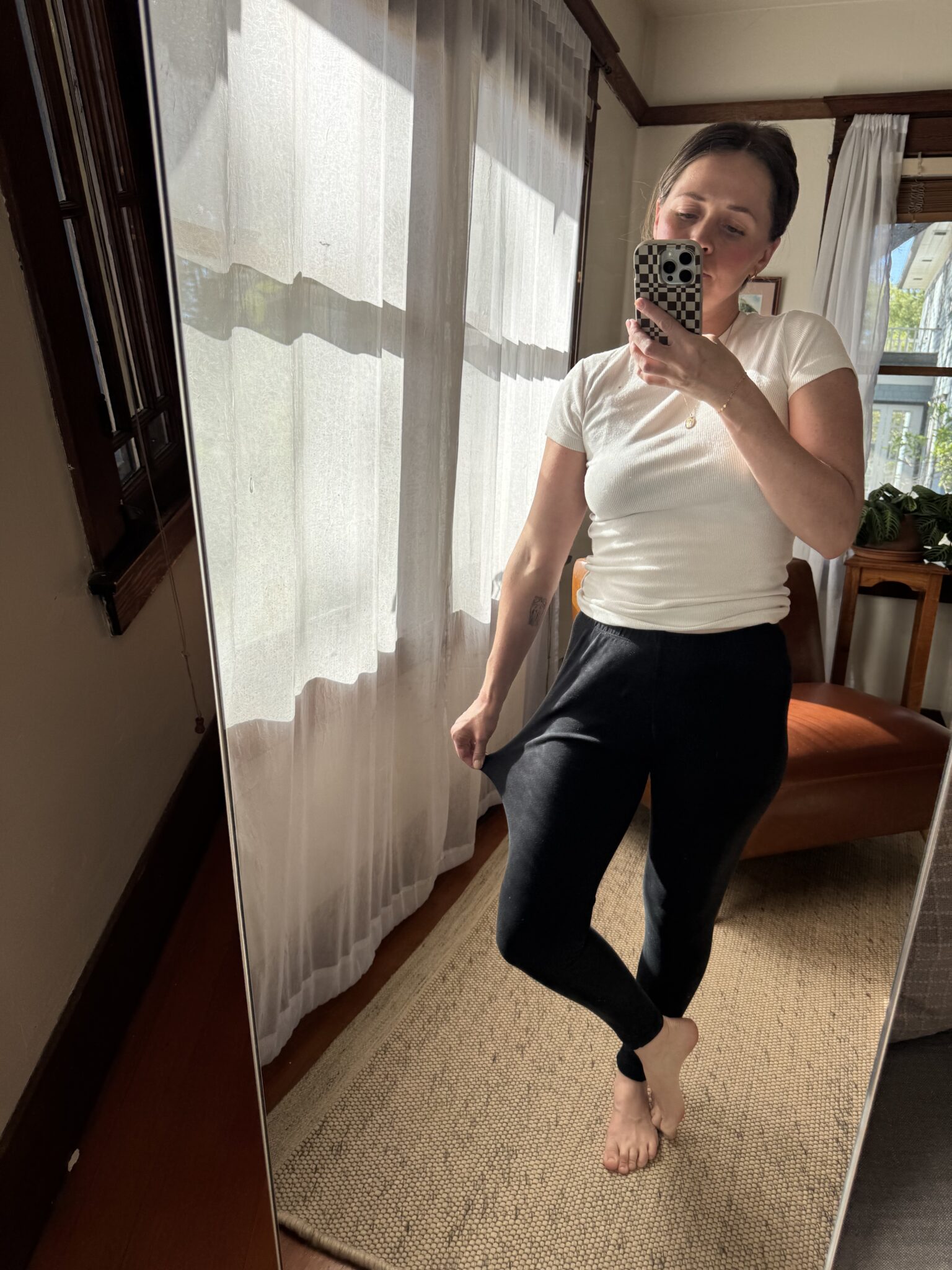 A person takes a mirror selfie in a well-lit room, wearing a white t-shirt and black pants, standing on a rug with sheer curtains in the background.