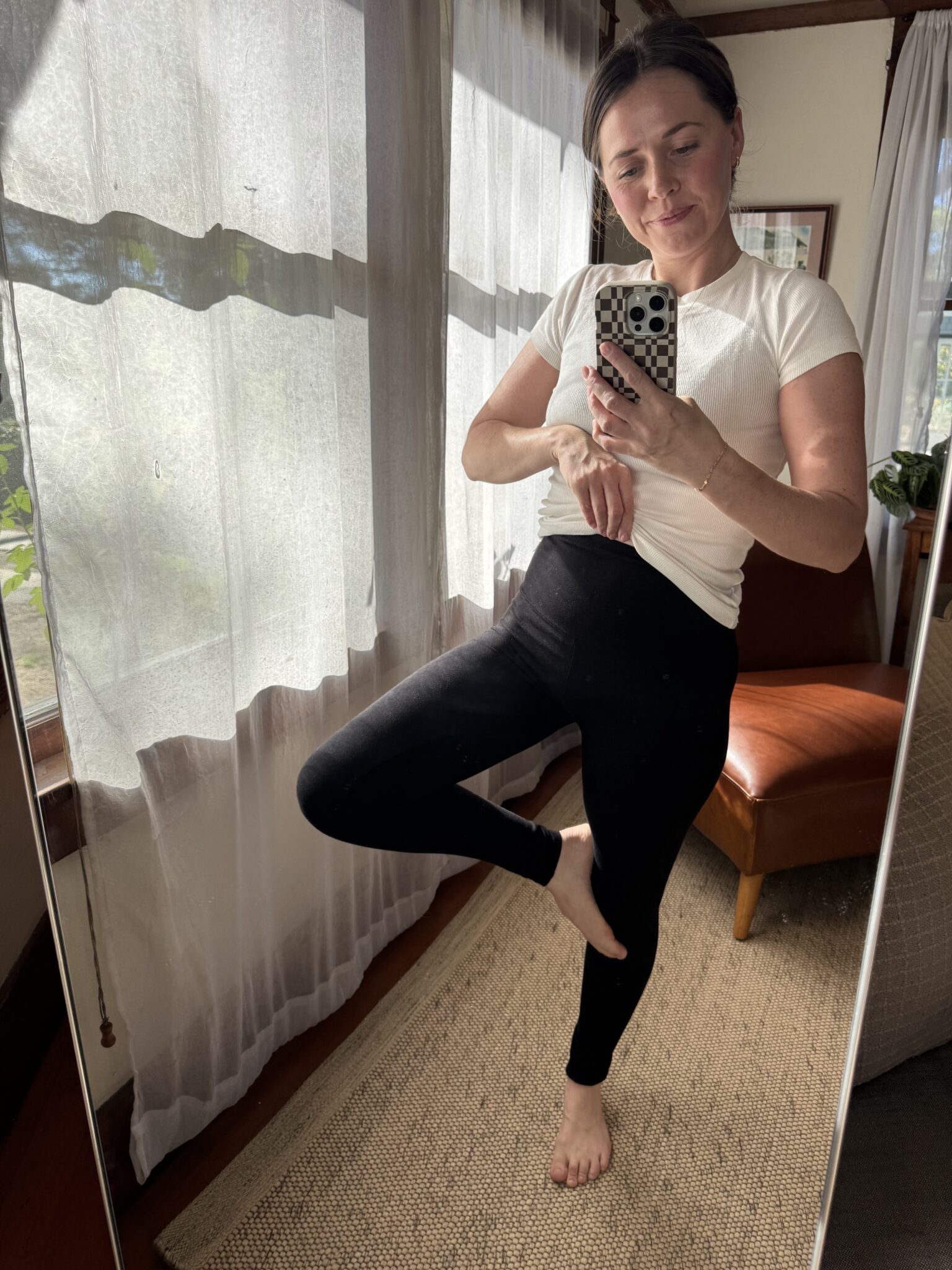 A woman in a white shirt and black leggings takes a mirror selfie, standing barefoot indoors with one leg bent, near a window with sheer curtains.