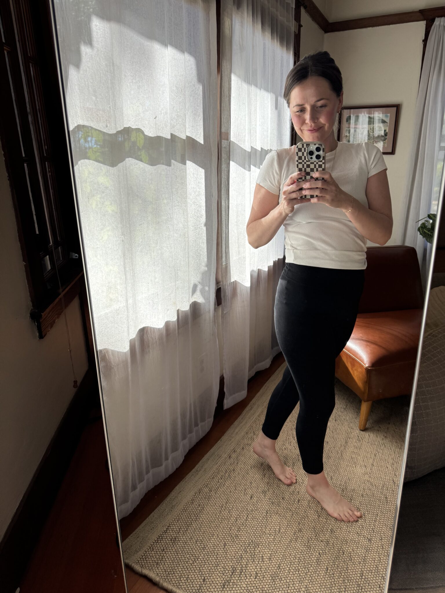 A woman stands barefoot on a rug, taking a mirror selfie in a bright room with sheer curtains and a brown chair.