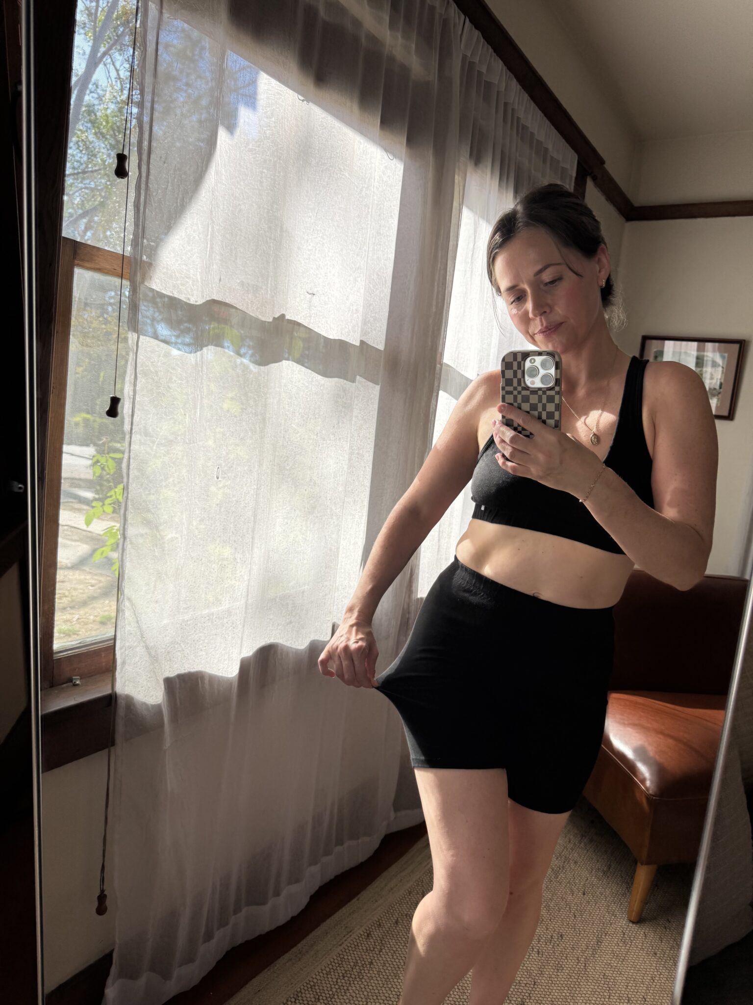 Woman stands indoors taking a mirror selfie, wearing a black sports bra and shorts, gently pulling at the waistband. Sunlight filters through sheer curtains behind her.