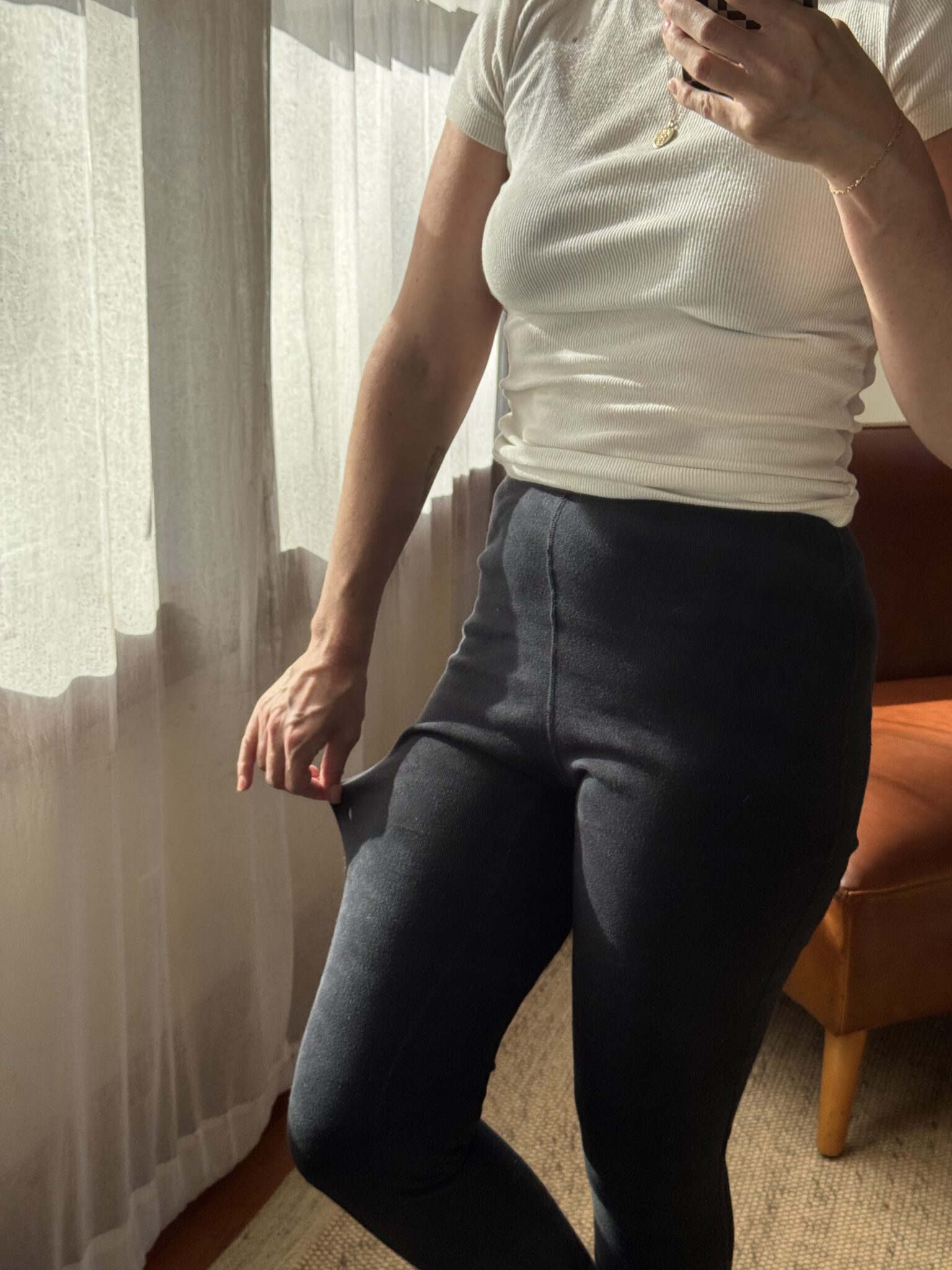 Person wearing a white ribbed short-sleeve top and dark high-waisted pants, standing indoors near a window, holding the side of their pants to show extra room.