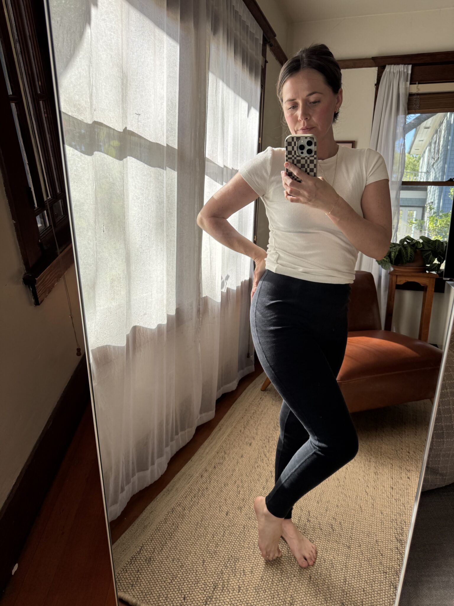 A person stands barefoot in a sunlit room, posing for a mirror selfie. They are wearing a white t-shirt and black pants. Light filters through sheer curtains in the background.