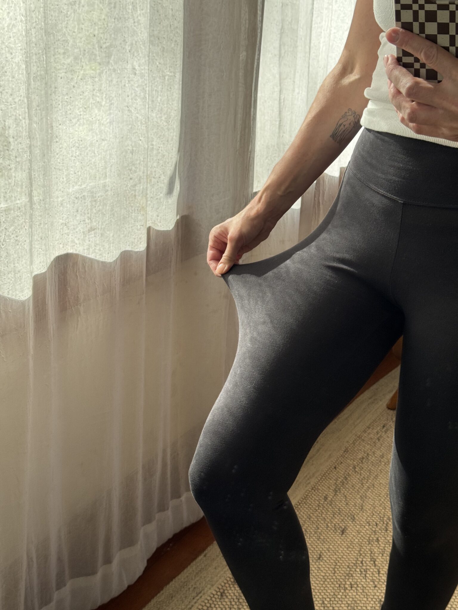 Person standing indoors, wearing dark leggings and a light top, pulling at the waistband of the leggings while taking a mirror selfie near a window with sheer curtains.