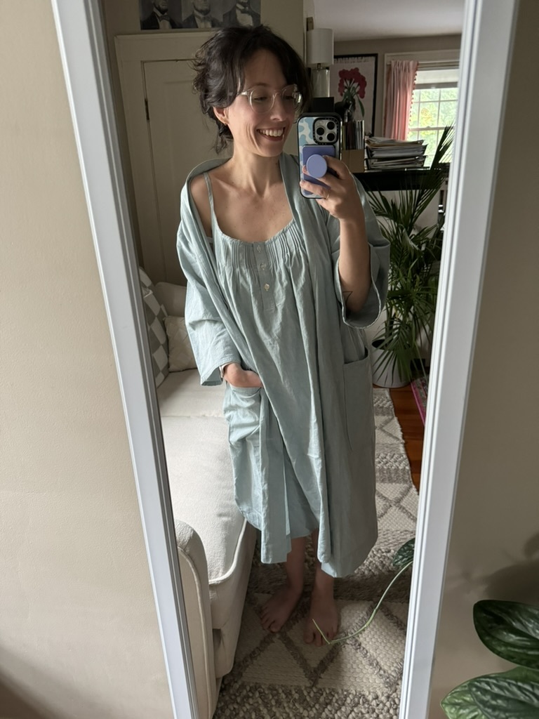 Person taking a mirror selfie indoors, wearing glasses and a light blue dress with a robe. There are plants, a couch, and framed art in the room background.