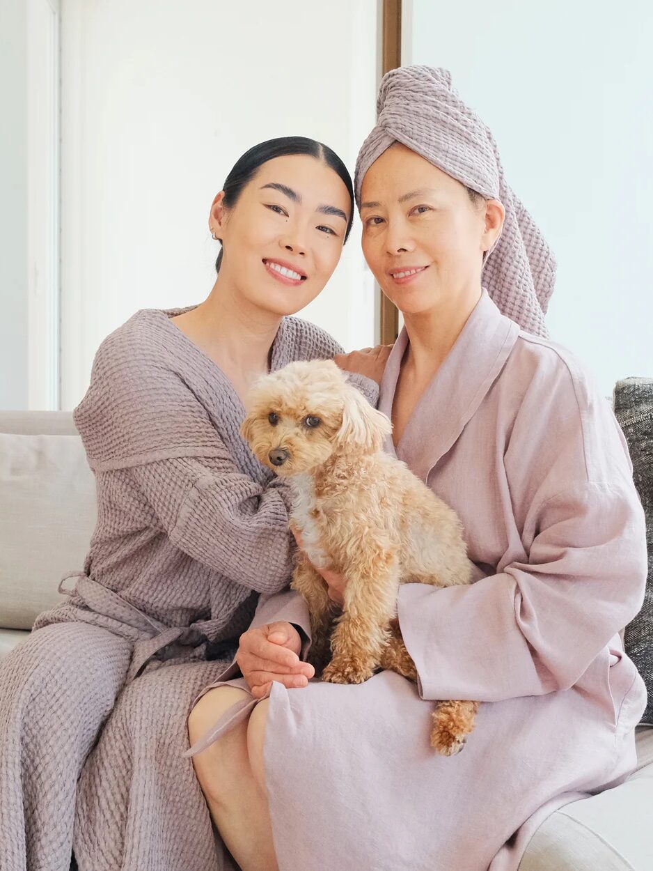 Two women in matching robes sit on a couch with a small, light-brown dog. One woman has a towel wrapped around her head. All three look toward the camera.