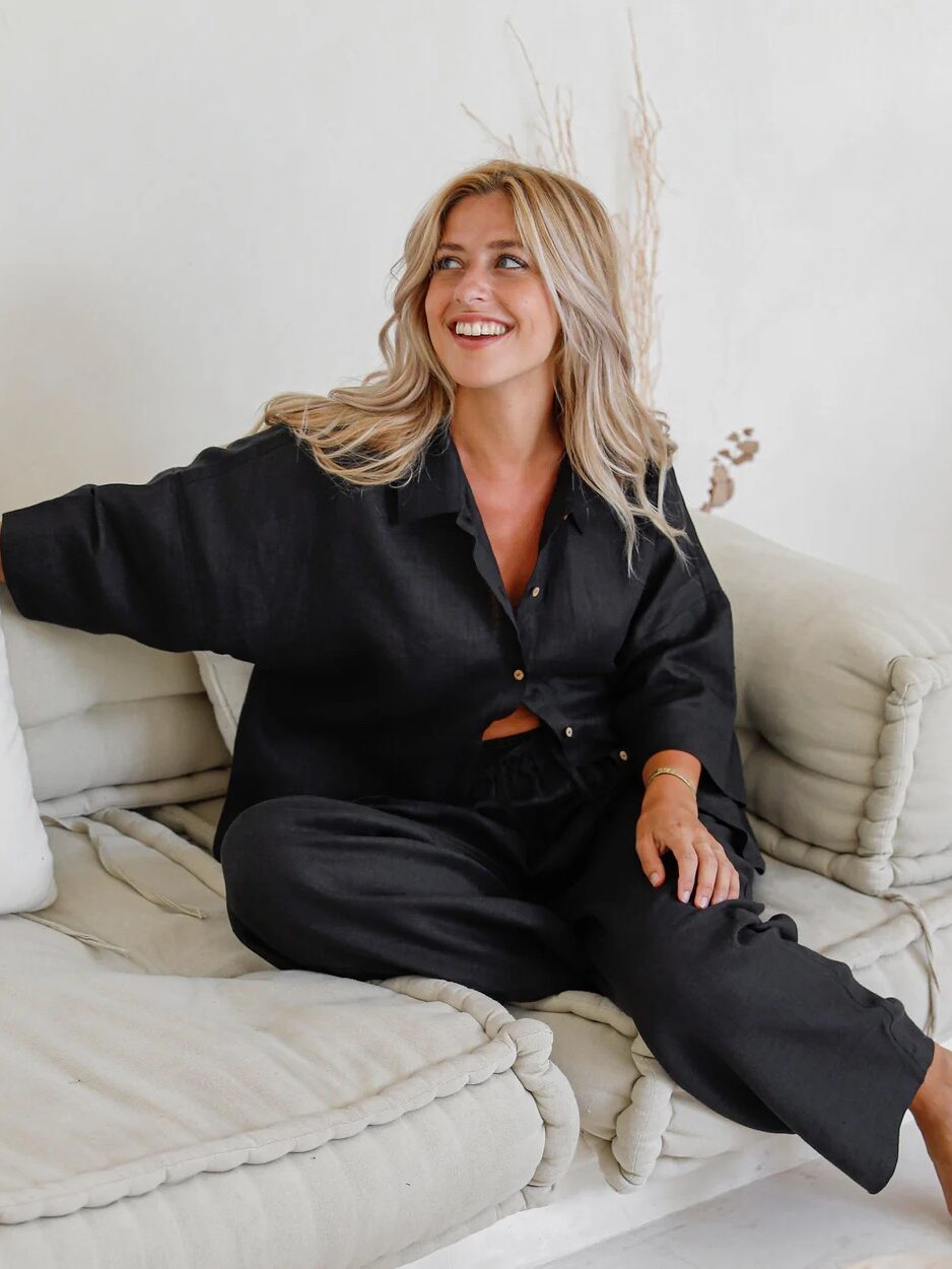 A woman with blonde hair sits on a beige cushioned sofa, wearing a loose-fitting black button-up shirt and matching pants, smiling and looking to the side.