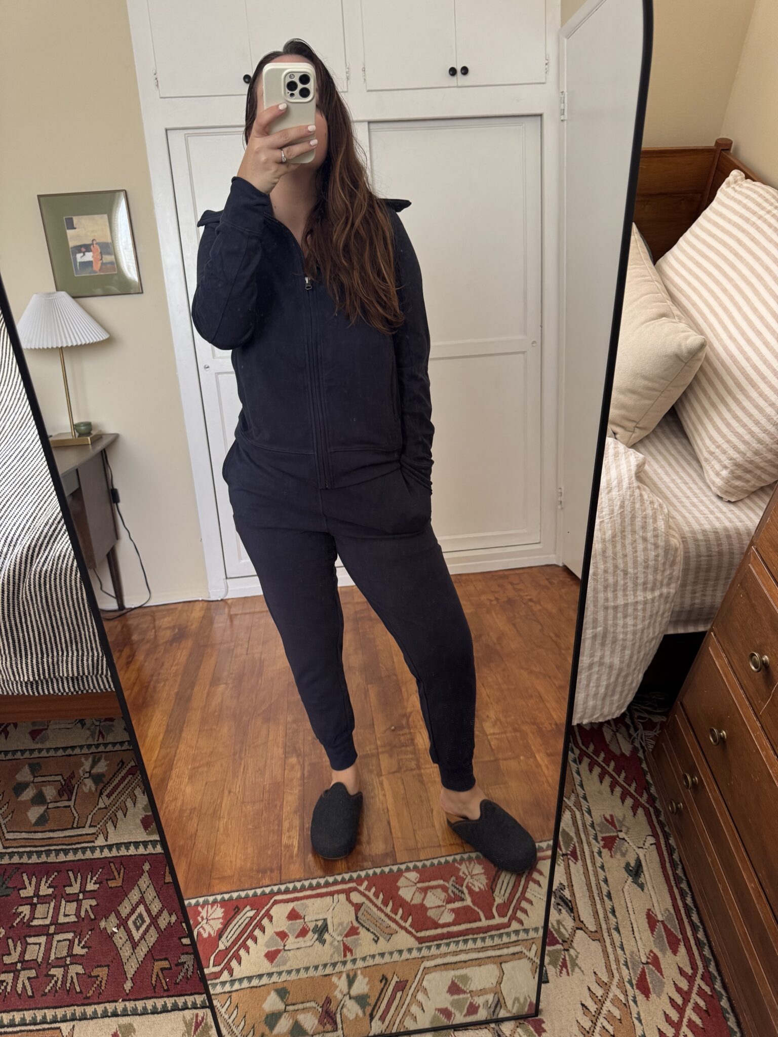 A person in a dark tracksuit and slippers stands in front of a mirror taking a selfie in a bedroom with wooden floors and patterned rugs.