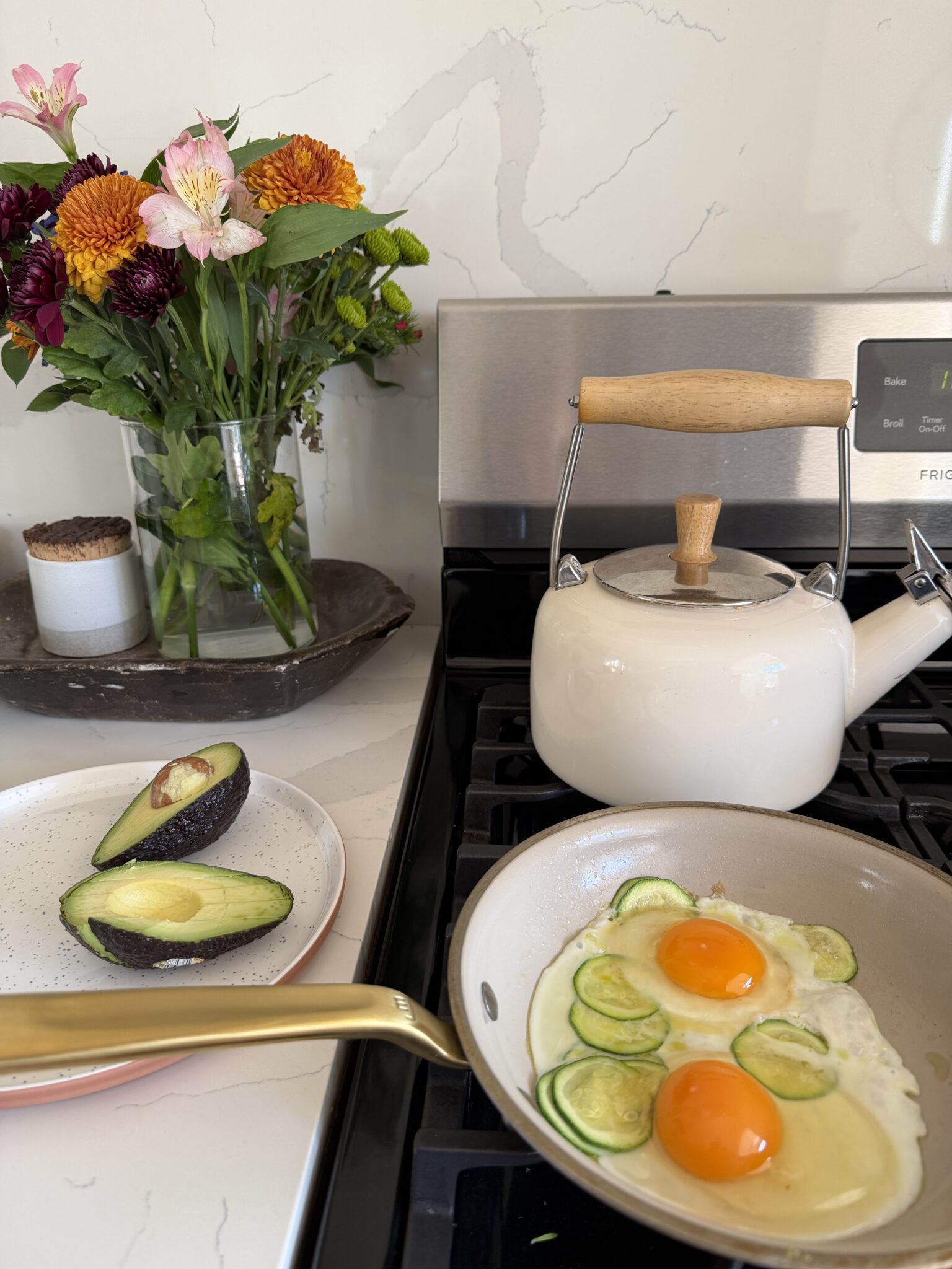 A frying pan with eggs and zucchini cooks on a stove next to a white kettle, with a plate of sliced avocado and a vase of flowers nearby.