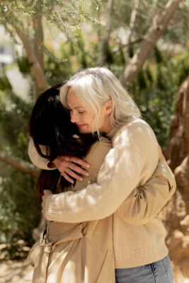 Two women are hugging outdoors in a sunlit area with trees in the background. The older woman has gray hair and is wearing a beige sweater; the younger woman has dark hair and a tan coat.