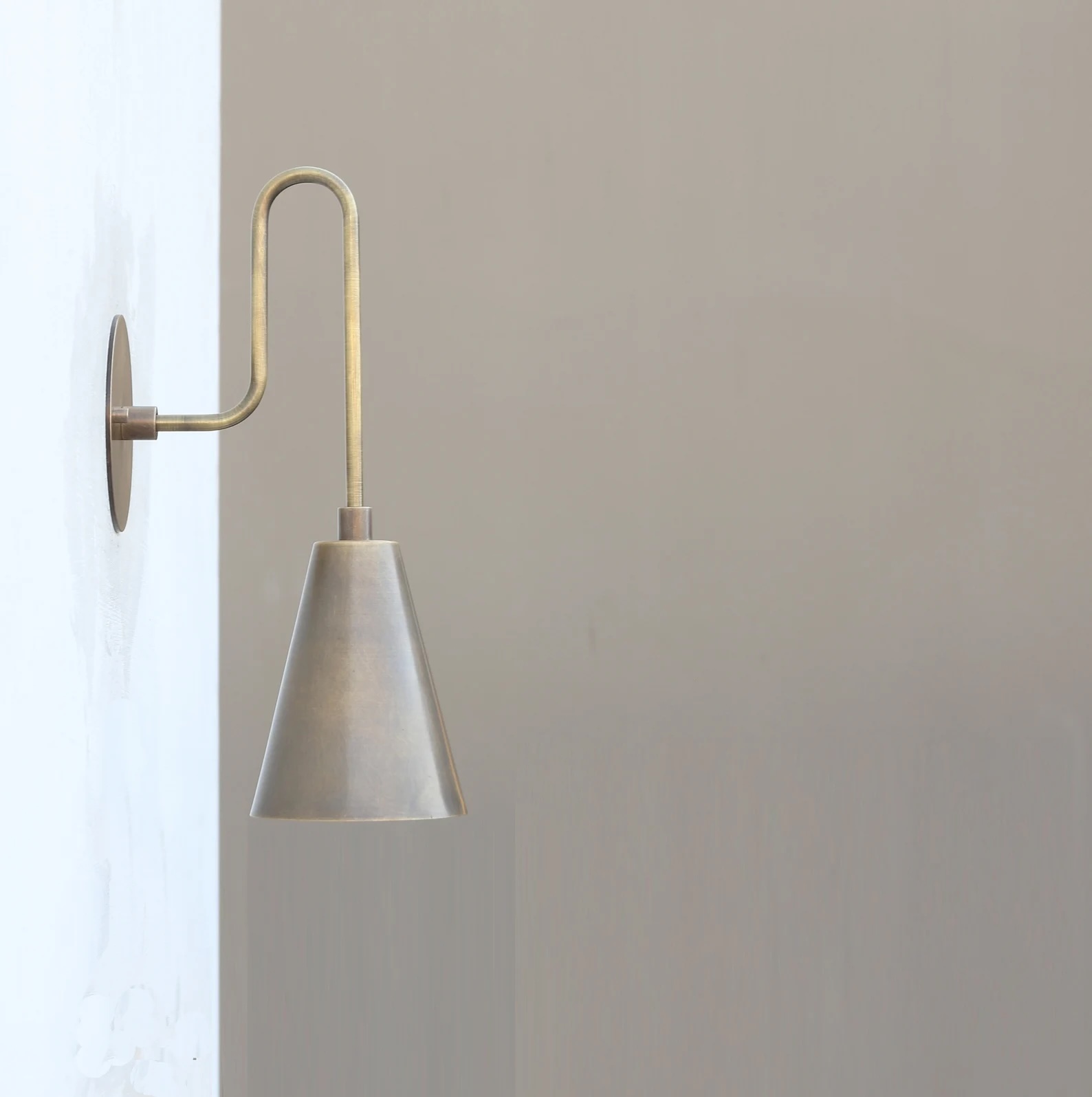 A brass wall sconce with a curved arm and conical shade is mounted on a plain, light-colored wall.
