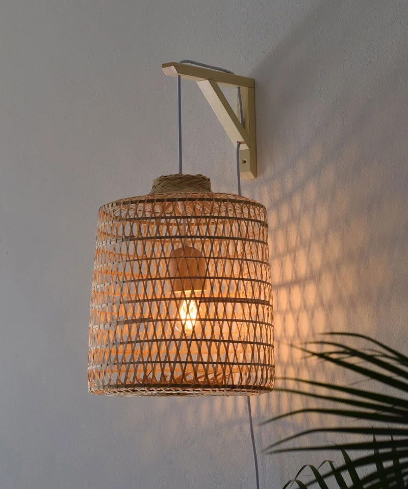 A woven rattan lampshade with a visible lightbulb is mounted on a wall bracket, casting a patterned shadow on the wall. A plant is partially visible in the lower right corner.