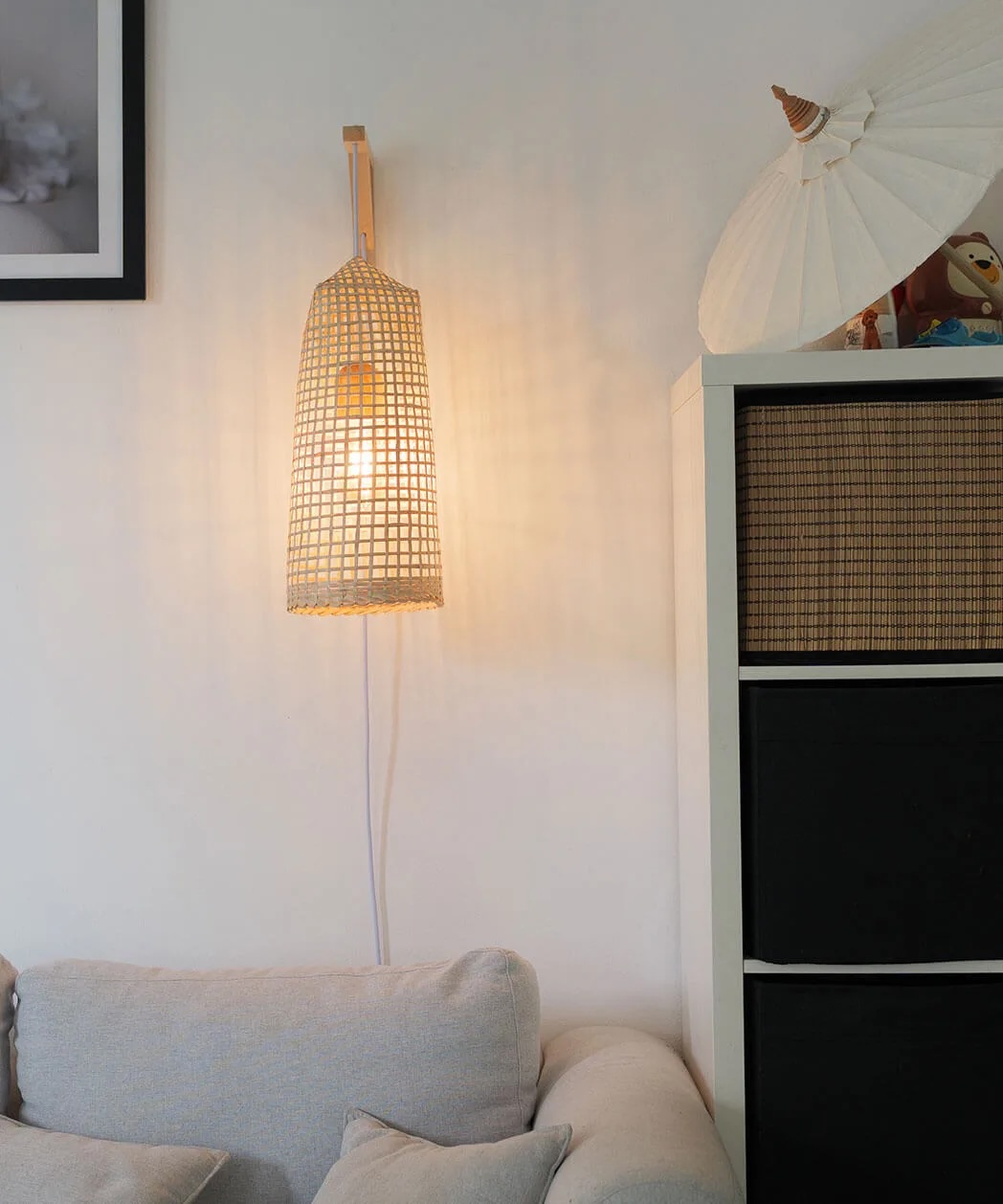 A woven lampshade wall light is illuminated above a couch next to a white shelving unit with baskets and an Asian-style paper umbrella on top.