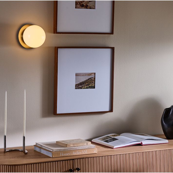 A wooden sideboard with books, an open magazine, and two tall candles sits below two framed photos and a round wall light on a beige wall.