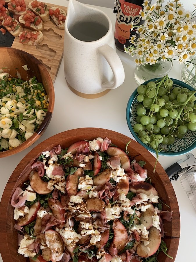 A table with a peach and prosciutto salad, a pasta dish, bruschetta, green grapes, a pitcher, and a bottle. Flowers are also on the table.