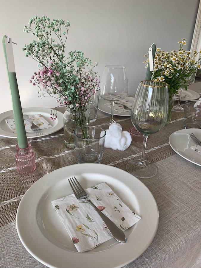 Elegant table setting with white plates, floral napkins, green candles, wine glasses, and small flower arrangements on a burlap runner.
