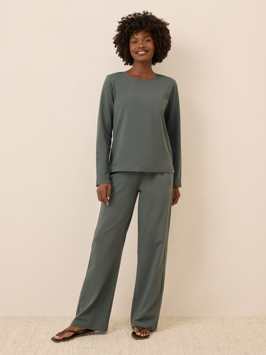 A person stands against a plain beige wall, wearing a matching long-sleeve top and wide-leg pants in muted green, paired with black sandals.
