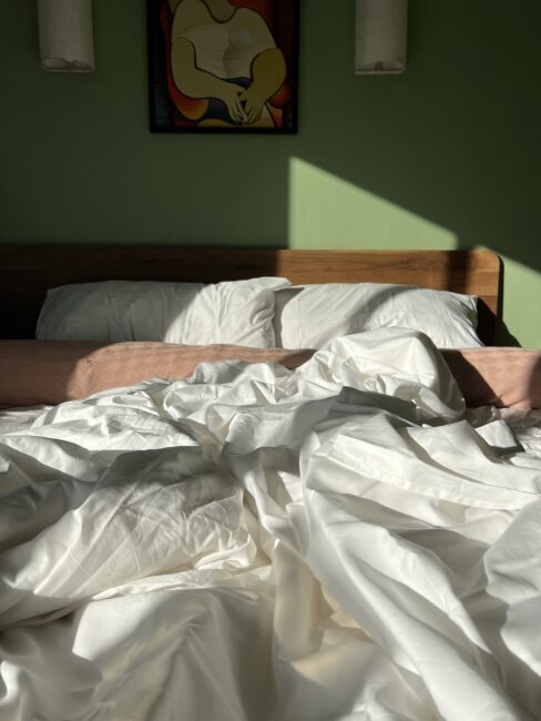 Unmade bed with white sheets in sunlight, two pillows against a wooden headboard, and a colorful abstract painting on the green wall above.