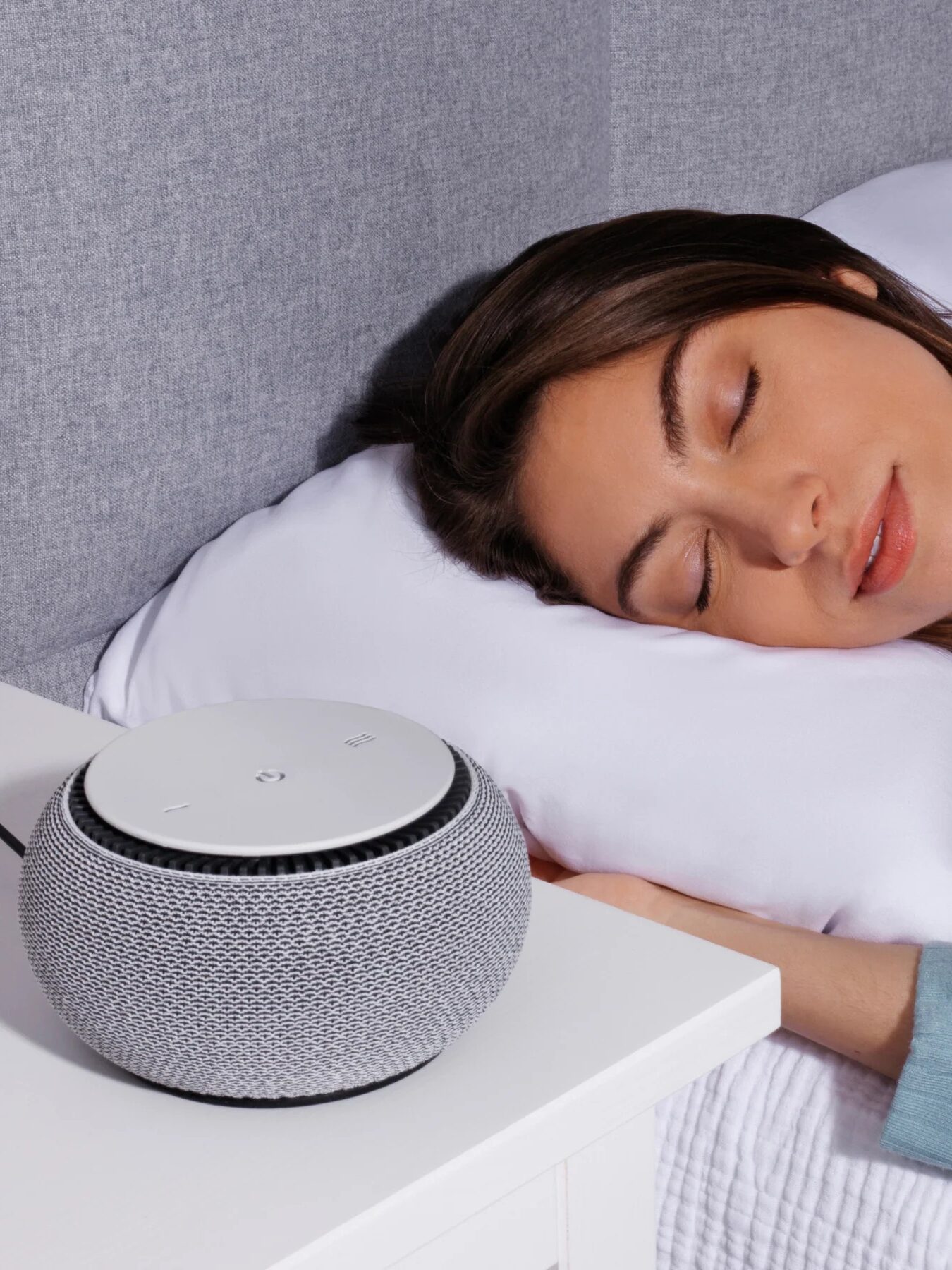 A woman sleeps on a white pillow next to a bedside table with a round, gray smart speaker placed on it.