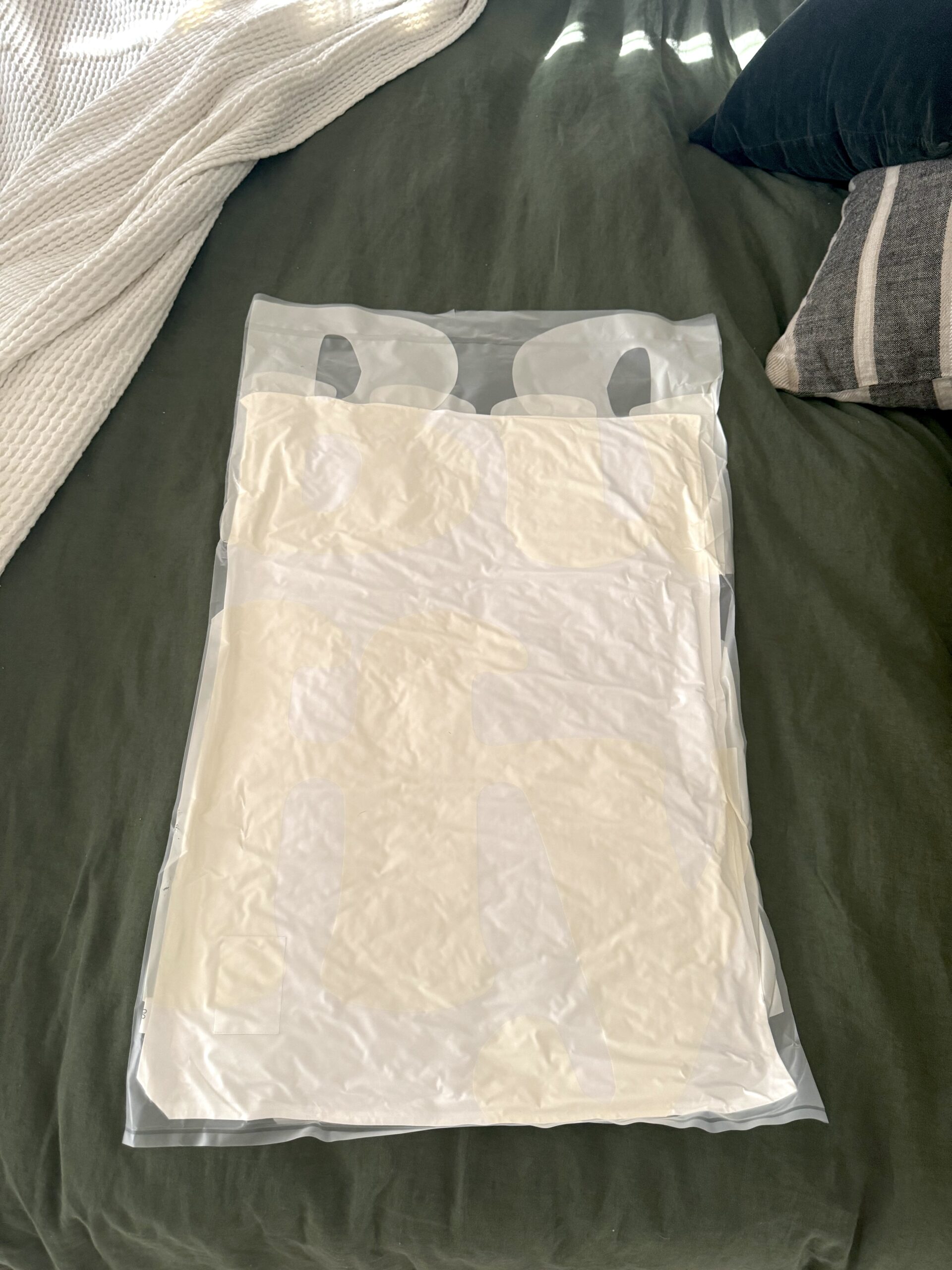 A vacuum-sealed plastic bag containing a folded white garment lies on a green bedspread next to a black pillow and a white blanket.