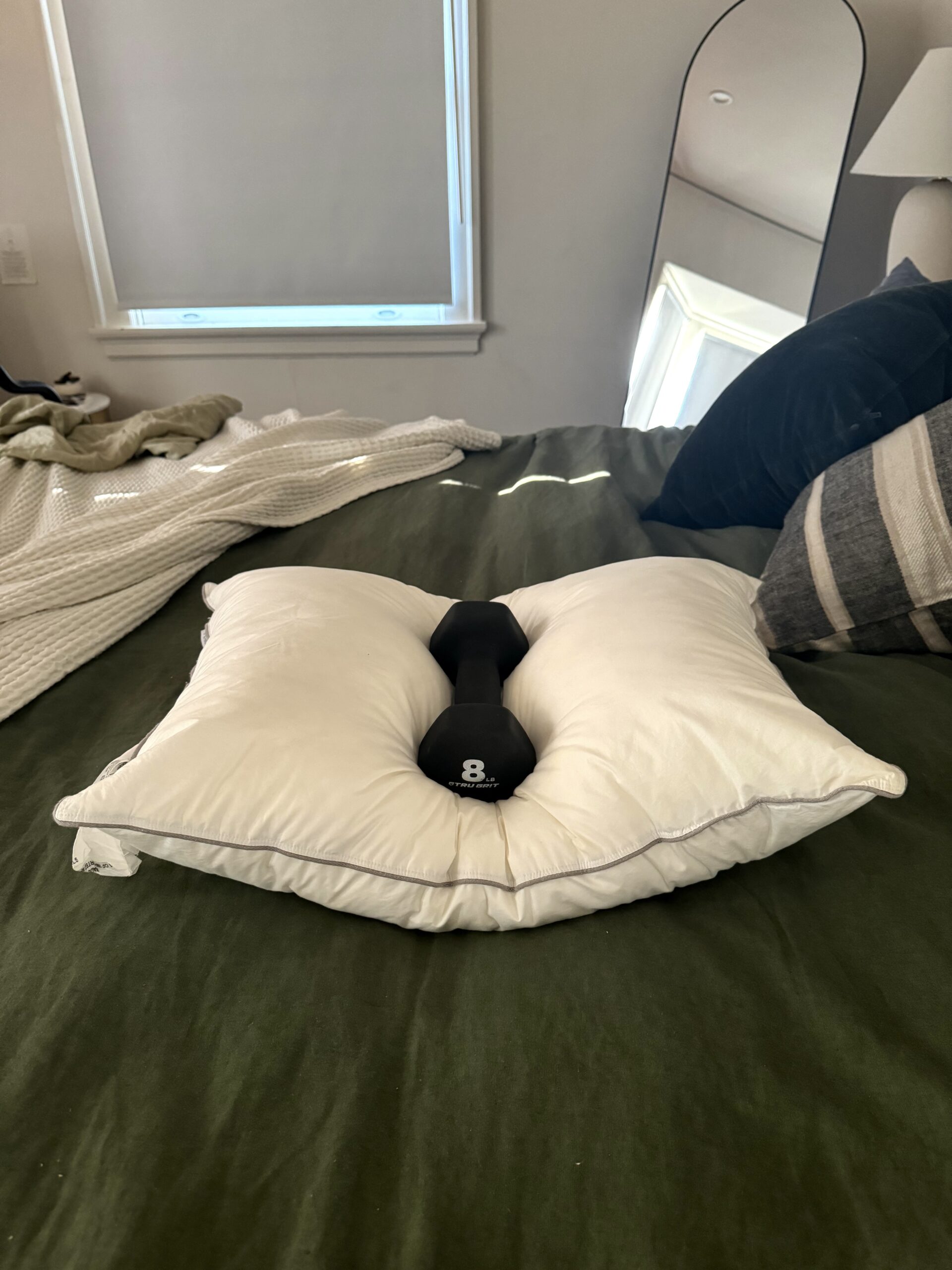 A white pillow with an 8-pound dumbbell placed on top rests on a green bedspread, with a mirror and another pillow in the background.