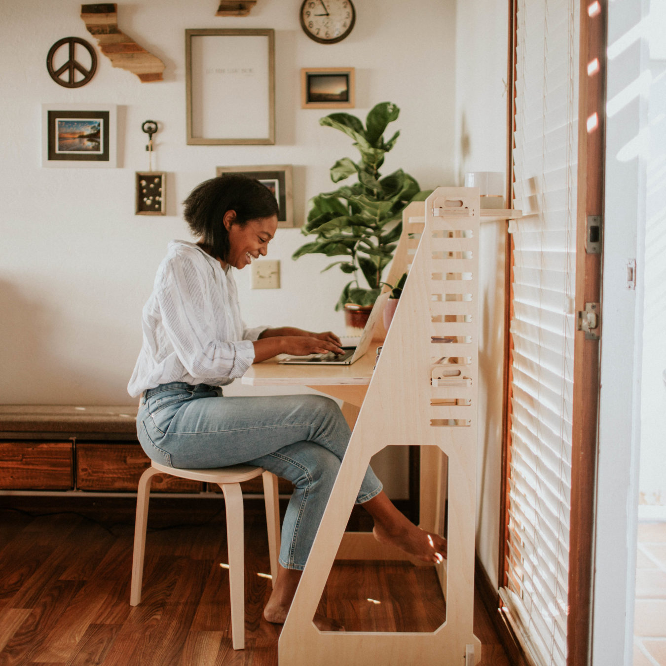 Woman sitting at a desk, using a laptop, in a well-lit room with wall art and plants.