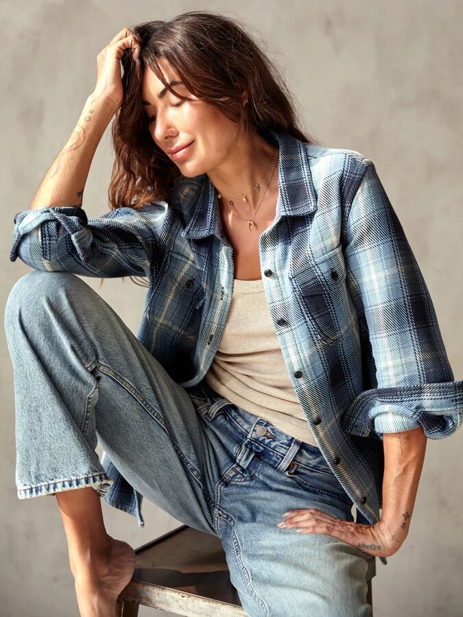 A person sitting on a stool, wearing a blue plaid shirt, jeans, and a beige top, smiling with eyes closed, hand touching their hair.