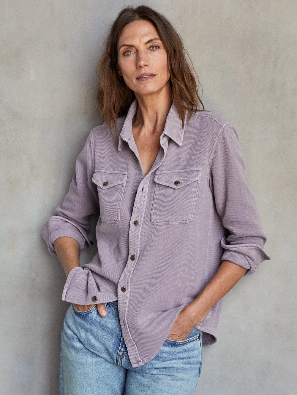 A person with shoulder-length hair wears a lavender button-up shirt and blue jeans, posing against a plain wall with hands in pockets.