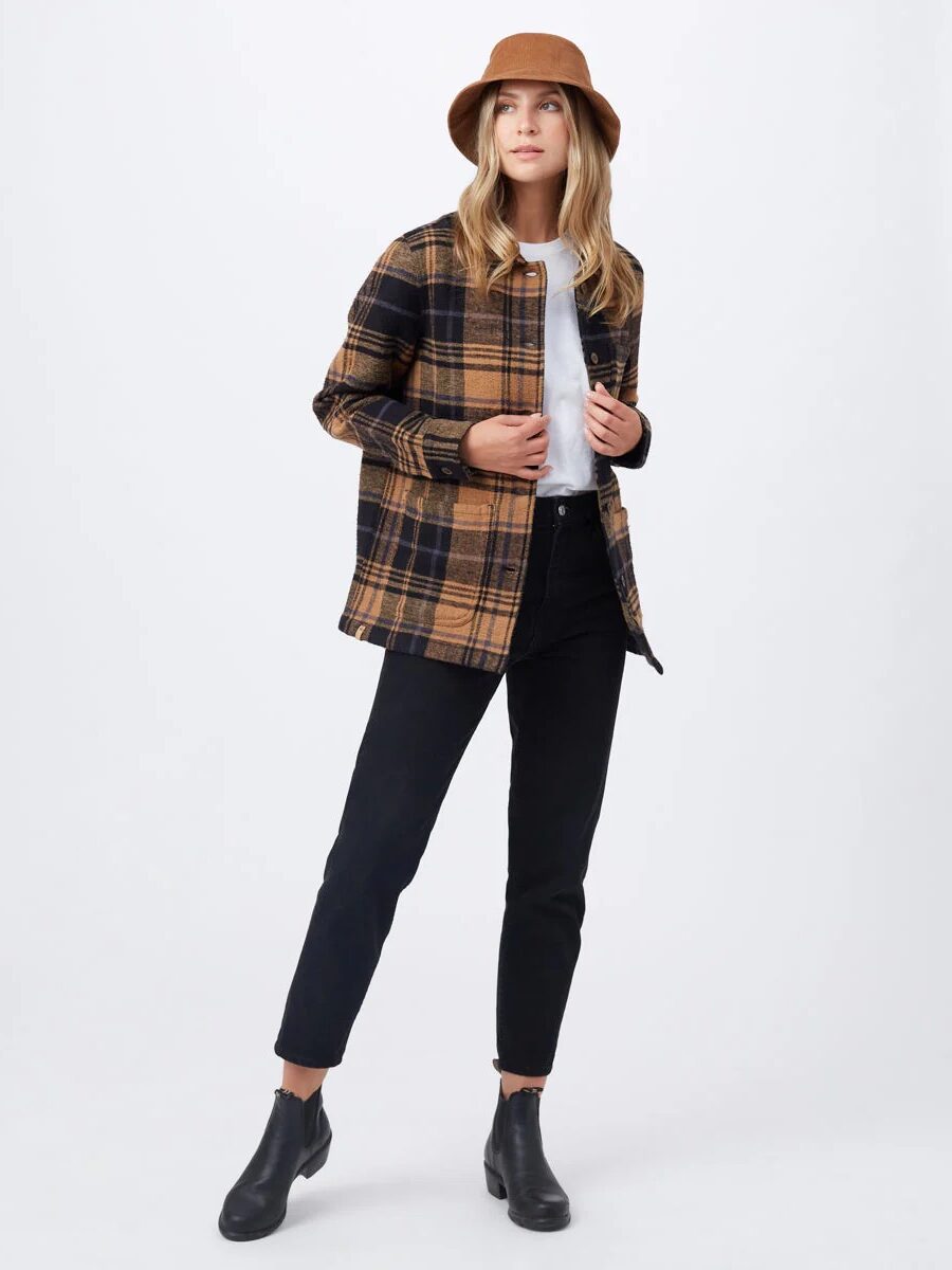 Person wearing a brown hat, plaid shirt, white top, black jeans, and black boots stands against a plain background.