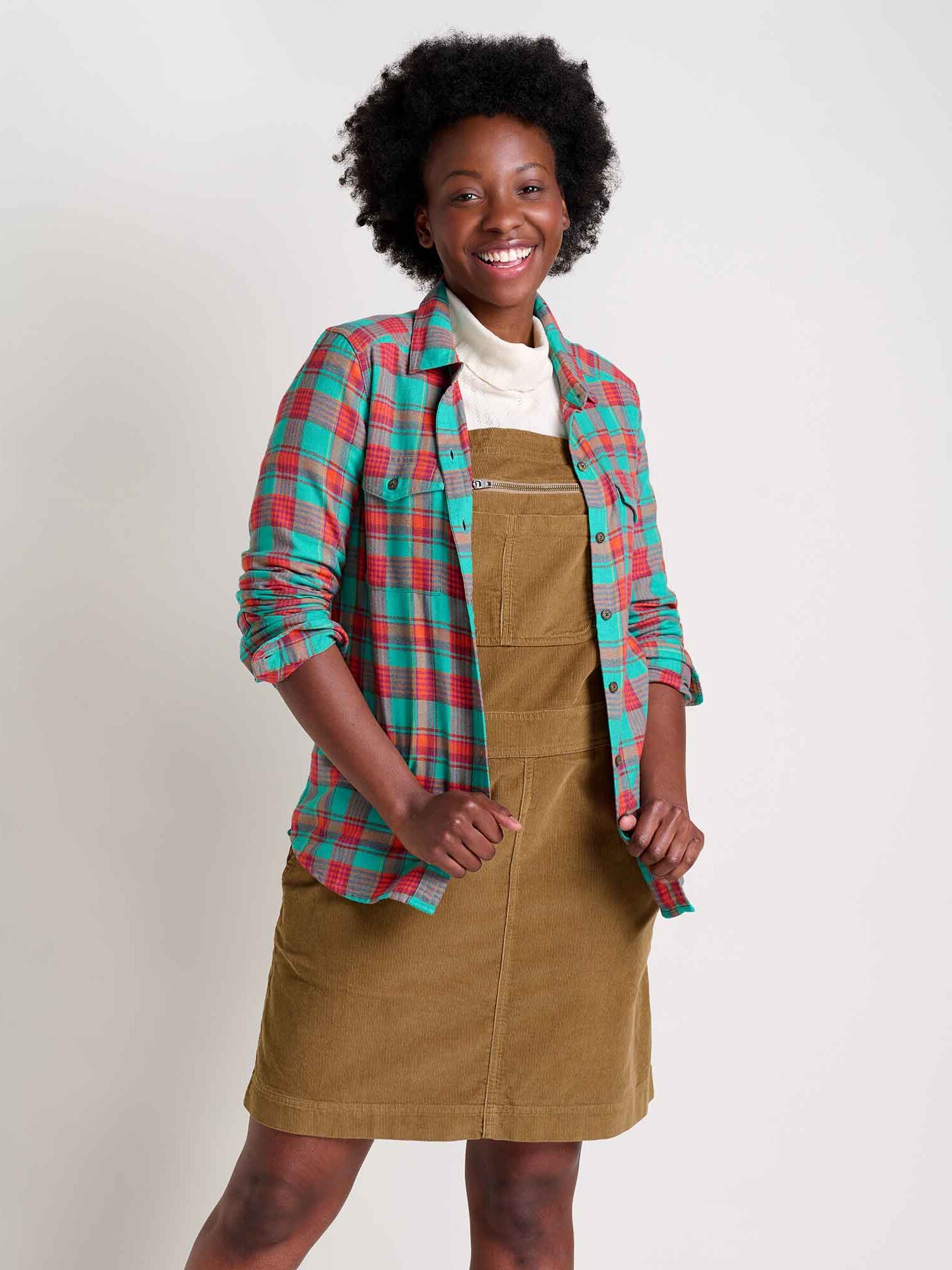 A person smiling, wearing a green and red plaid shirt over a tan dress, standing against a plain background.