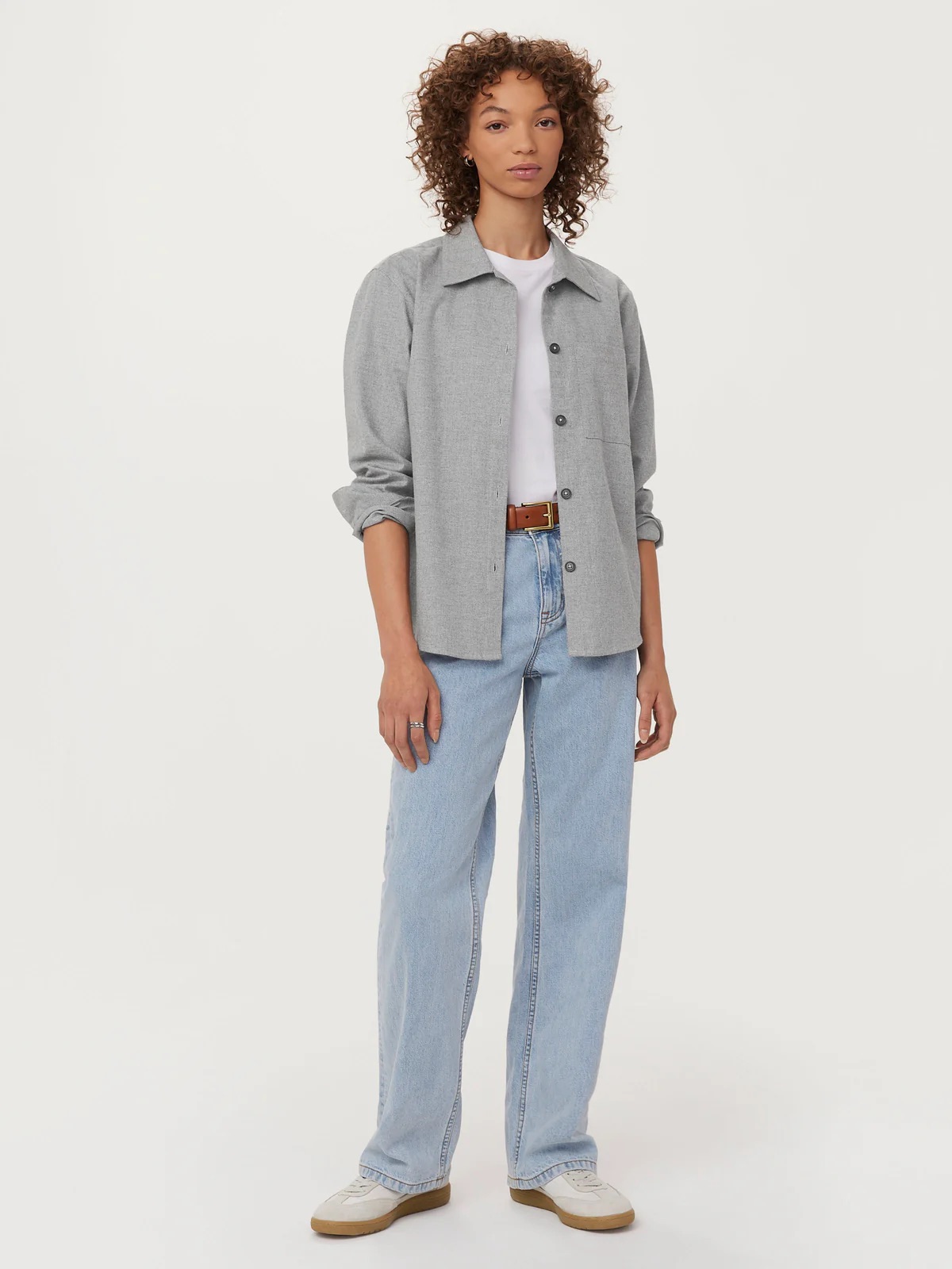 A person with curly hair wears a gray shirt, white t-shirt, light blue jeans, and white sneakers. They stand against a plain white background.