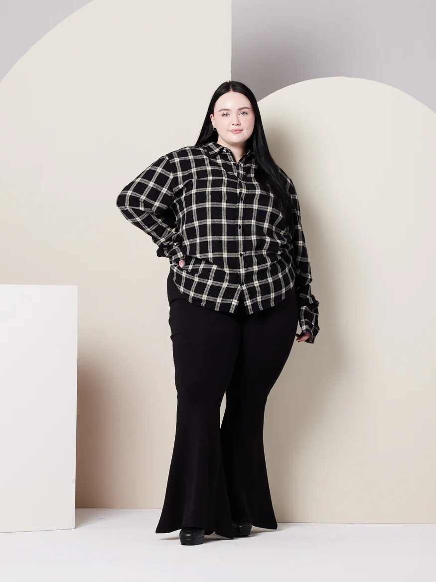 Person in a black and white plaid shirt and black pants stands against a neutral background with geometric shapes.