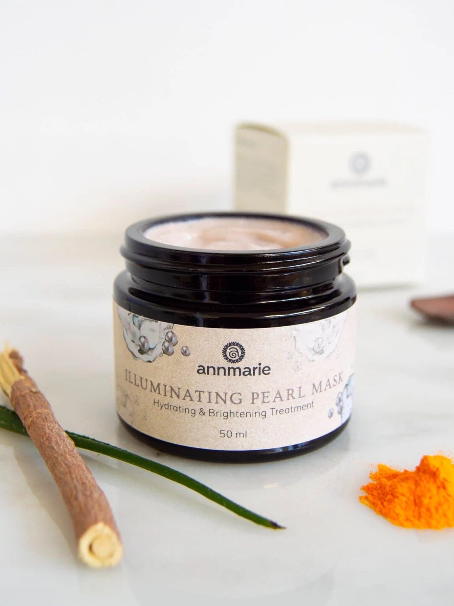 A jar of Annmarie Illuminating Pearl Mask with a brush, a pile of yellow powder, and a green leaf on a white surface; the product box is in the background.