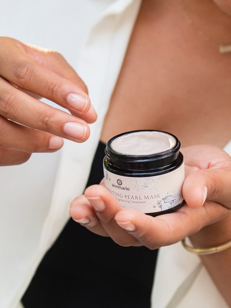A person holds an open jar of Leahlani Mermaid Mask in one hand and uses fingers of the other hand to scoop out some product, wearing a white shirt and gold bracelet.