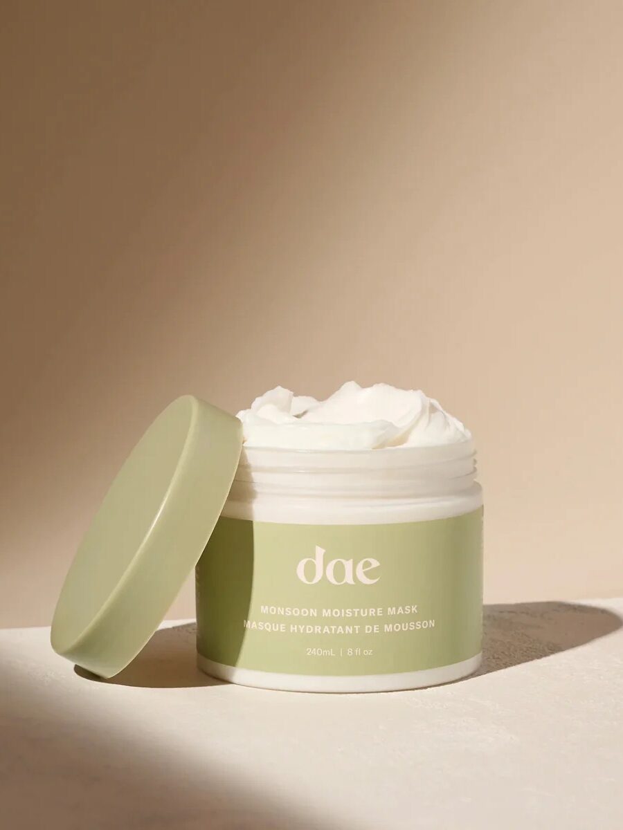 A jar of Dae Monsoon Moisture Mask with the lid off, showing the creamy white product inside, is placed on a neutral beige surface.