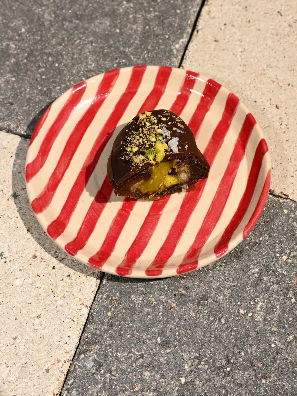 A chocolate-covered dessert with a yellow filling and crushed pistachios on top, served on a red and white striped plate placed on a tiled surface.