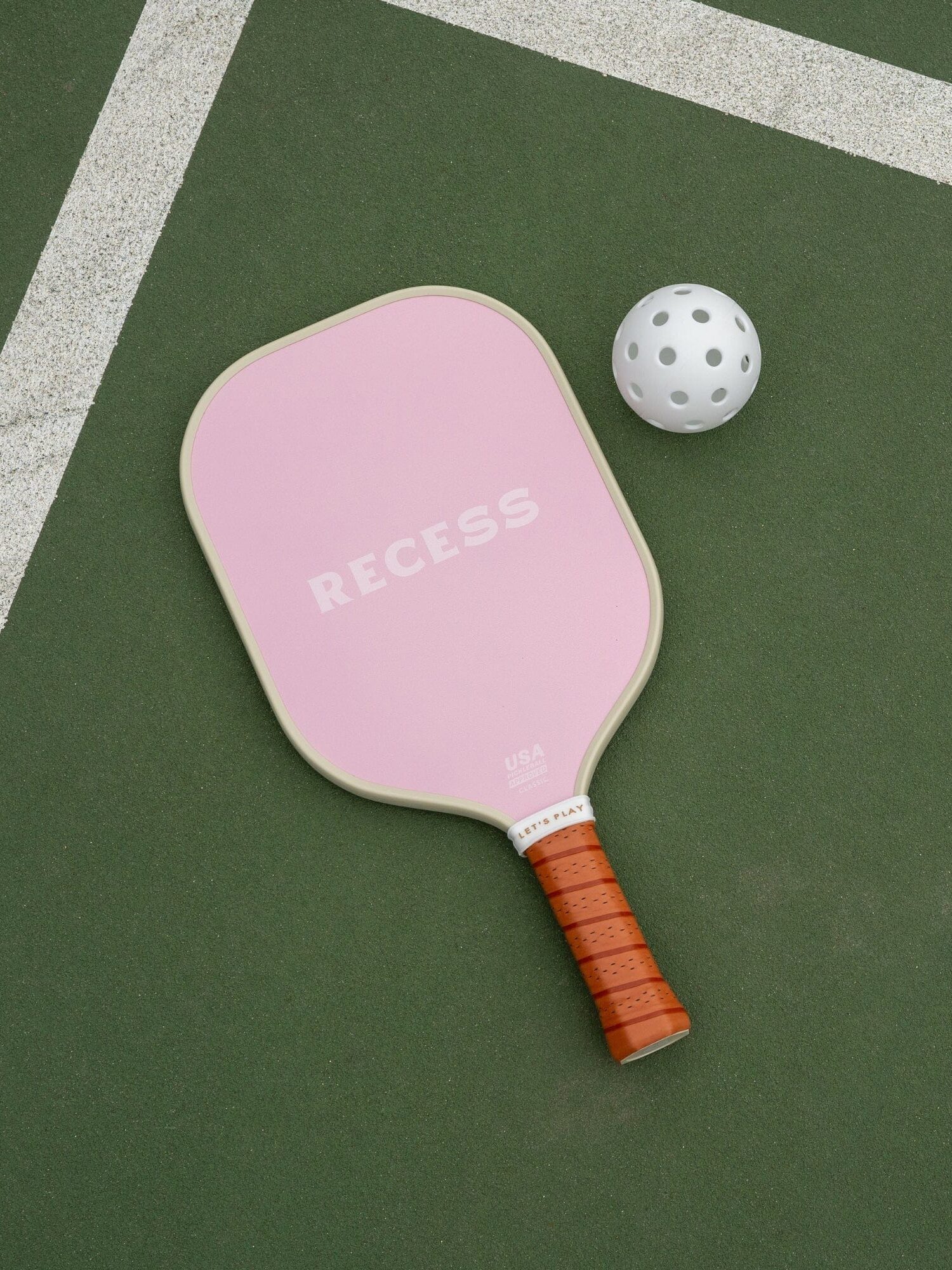 A pink pickleball paddle labeled "Recess" and a white perforated ball lie on a green court near white boundary lines.