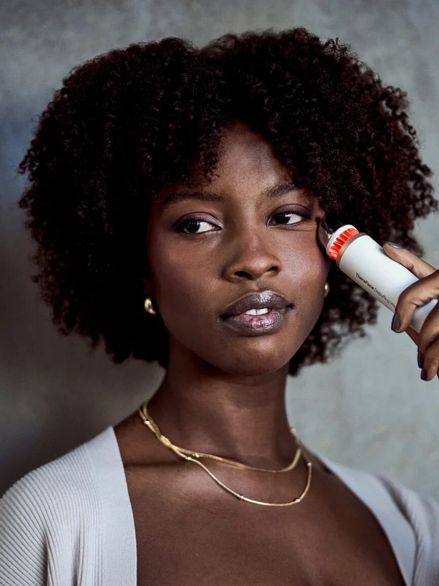 A woman holds a white container with a red lid to the side of her face against a neutral background.