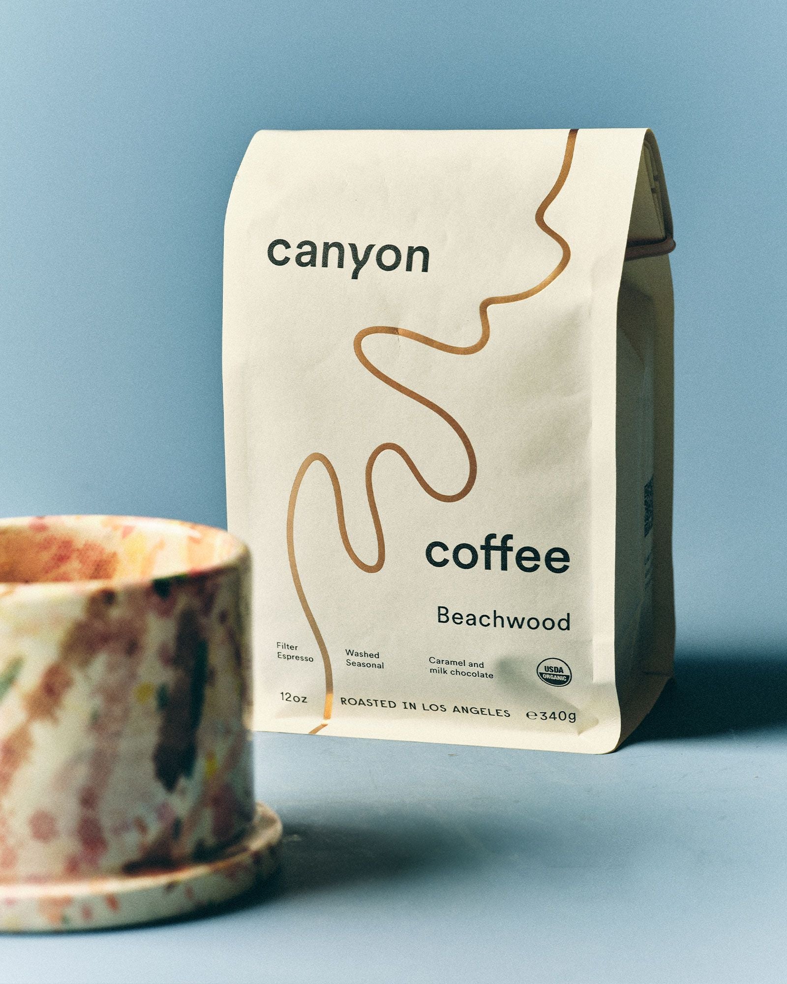 A speckled ceramic mug sits in front of a beige Canyon Coffee Beachwood bag against a light blue background.