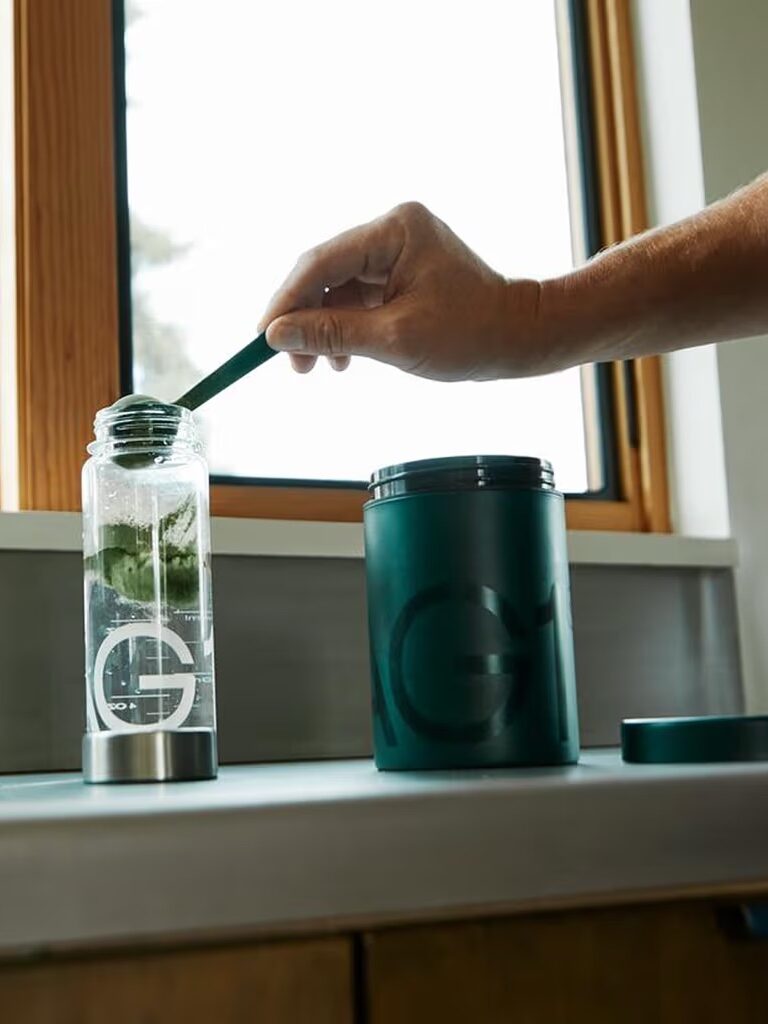 A person stirs green powder into a clear water bottle next to a matching green container on a kitchen counter by a window.