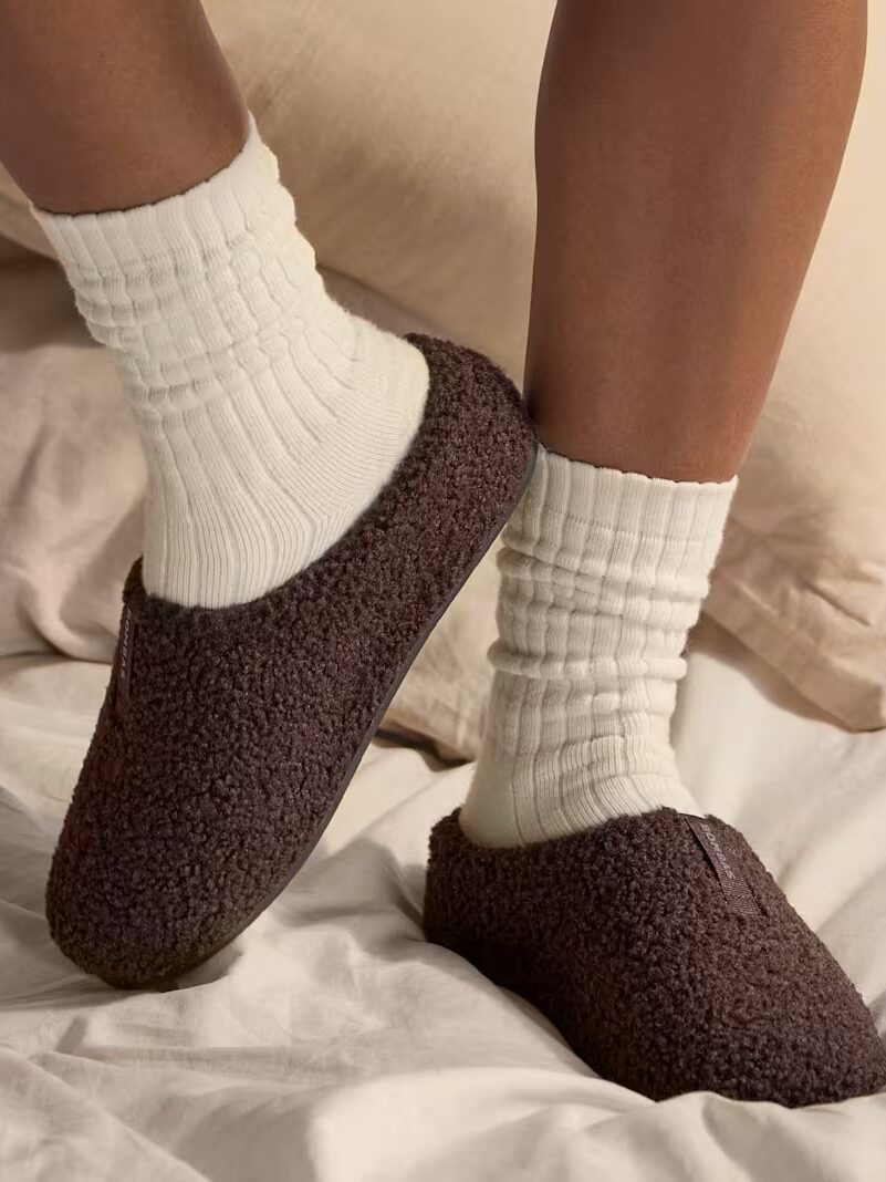 A person wearing white ribbed socks and dark brown fleece slippers stands on a bed with light-colored bedding.