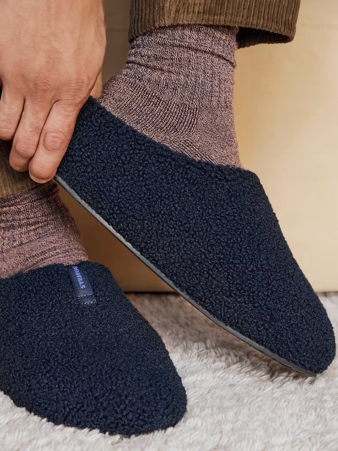 A person wearing brown socks and dark blue textured slippers sits on a light-colored surface, adjusting the left slipper with their hand.